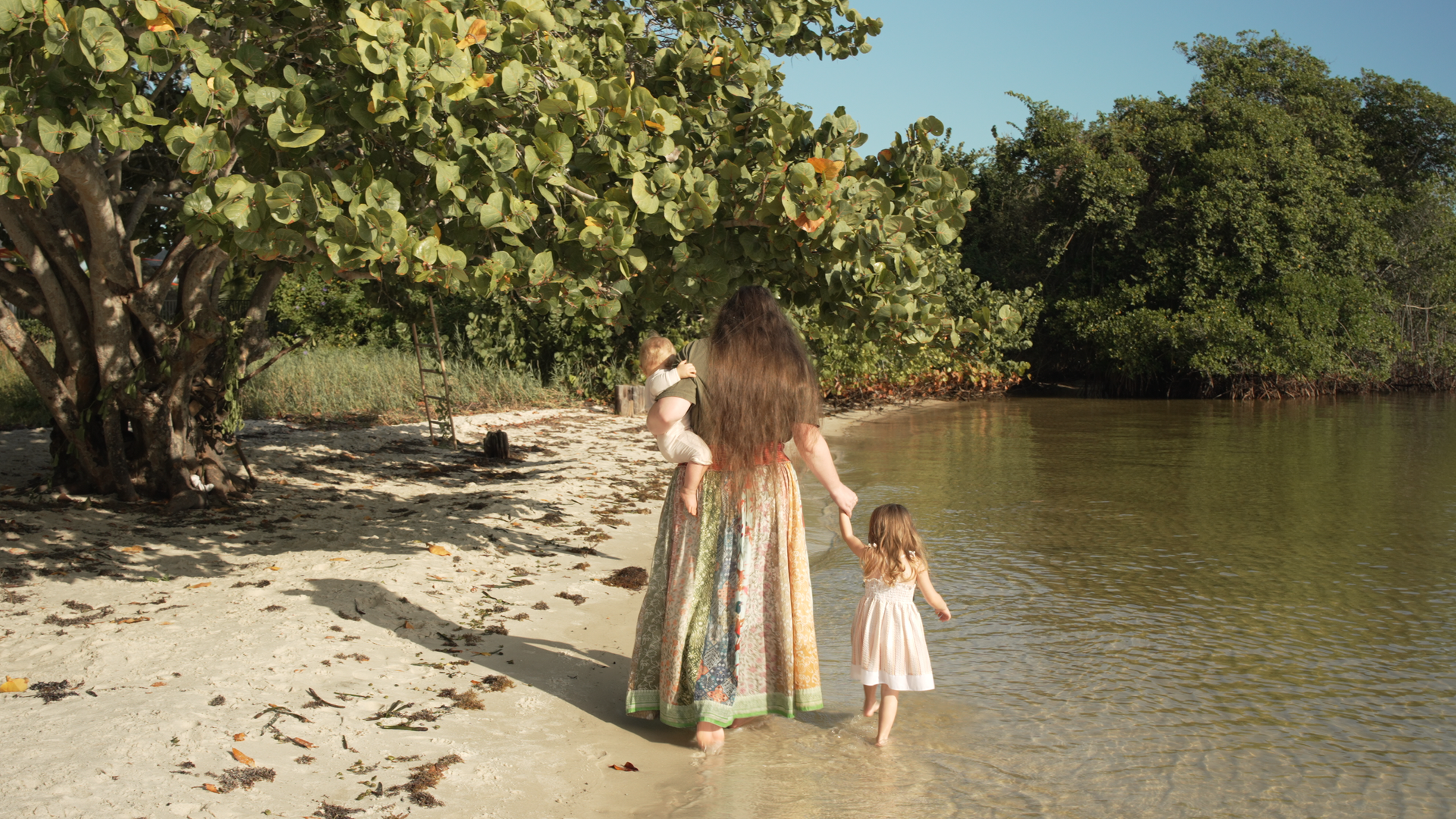 Founder of Earthspun Loom walking along the shoreline holding a baby and a toddler, wearing a cotton dress, reflecting slow living and family life