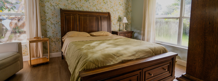 Organic cotton bedding styled on a master bed in a cozy, light-filled bedroom with dark wood furniture, floral wallpaper, and wood floors, reflecting natural materials and slow living